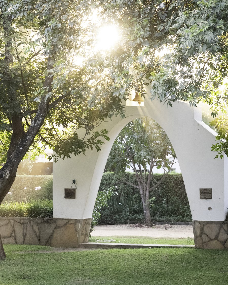 The white memorial arch and bell beneath the trees on the Musikili campus